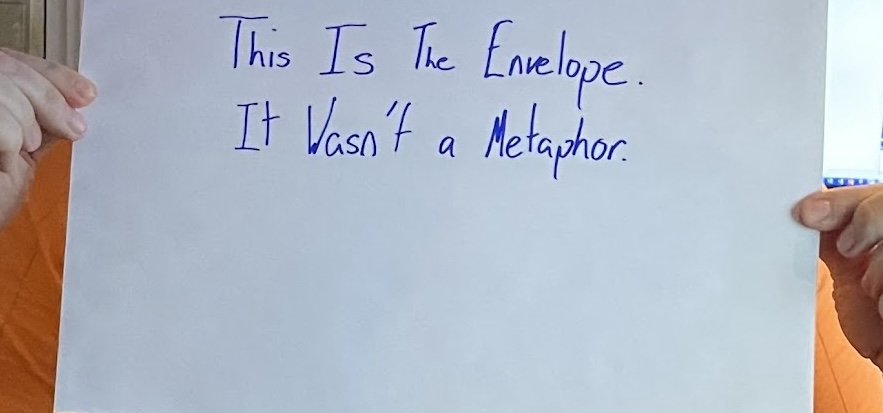 A severely zoomed in and cropped image of hands holding an envelope that reads "This is the envelope. It wasn't a metaphor."